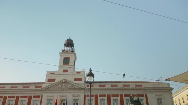 Low angle view of the historic Real Casa de Correos building in Puerta del Sol, Madrid, featuring the iconic clock tower and pigeons flying against a clear blue sky.