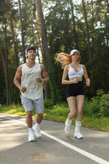 Fototapeta premium Fit young couple jogging together on a paved park road. Athletic man and redhead woman running in stylish sportswear during a sunny summer cardio workout in a pine forest setting.