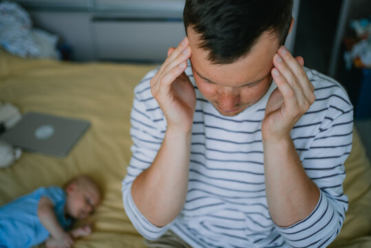 Frustrated father holding his temples and suffering from a headache, feeling overwhelmed by parental responsibilities next to his sleeping baby, representing paternal stress
