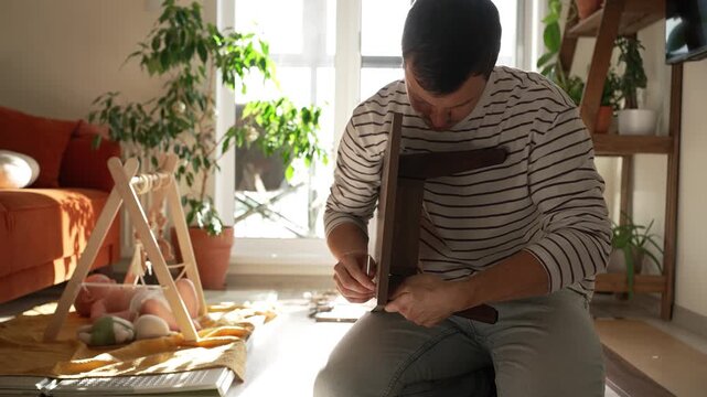 Young dad skillfully assembles a wooden chair on the floor while his infant child sleeps nearby, demonstrating a dedicated father's ability to balance household chores and childcare