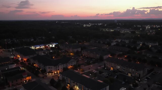 Pink and orange sunset glows above dense apartment rooftops in Cornerstone, Pennsylvania, warm porch lights and illuminated storefront dot sprawling suburban housing below. Aerial drone rising shot