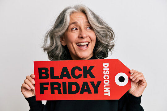 Excited mature woman with grey hair smiles and holds a red BLACK FRIDAY DISCOUNT tag, ready for shopping spree.
