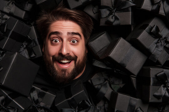 A man's ecstatic face emerges from a sea of black gift boxes, his wide eyes and open-mouthed smile conveying overwhelming joy and excitement.