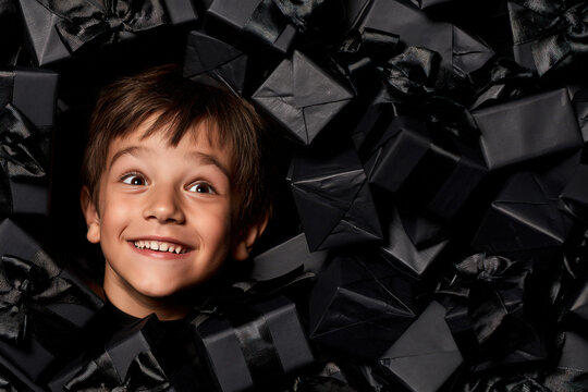 A young boy peeks out from a pile of black gift boxes, his eyes wide with excitement and a mischievous smile playing on his lips. The dark.