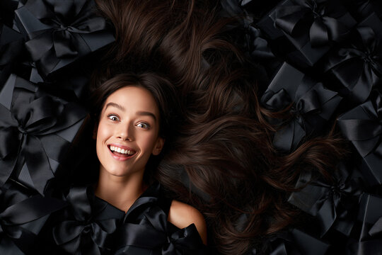 A joyful young woman with flowing dark hair smiles widely, lying amidst a bounty of luxurious black gift boxes, embodying the excitement of surprise and generosity.