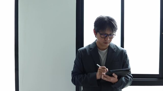 Asian businessman wearing suit and glasses writing on tablet with stylus pen near office window. Focused male professional taking digital notes, planning, and managing business project