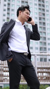 Asian businessman in suit talking on smartphone and texting outdoors. Low angle view optimized for mobile display, social stories, business connection, and mobile ads