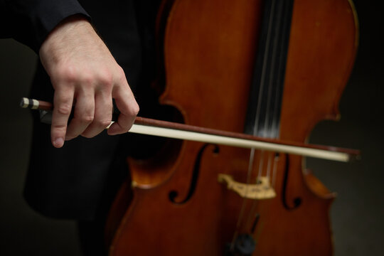 Caucasian young adult cellist hand holding bow playing acoustic cello in orchestral concert setting. Visual suitable for classical music education and cultural events promotion
