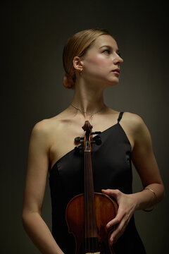 Vertical shot of young adult Caucasian woman holding acoustic violin, looking away. Classical music performer concept suitable for concert promotion and musical education marketing