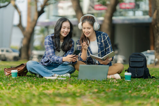 Two Asian female college students studying together and reading book on grass lawn in university campus.
