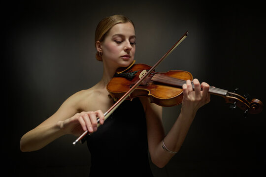 Young adult Caucasian female violinist playing classical violin with bow against studio background. Image suitable for music school advertising, concert promotions and arts education