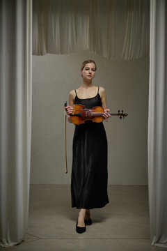 Vertical portrait of young adult Caucasian woman holding violin and bow standing on stage. Musician preparing for classical concert performance
