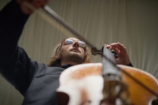 Low angle view of young adult Caucasian man playing cello instrument during classical music performance rehearsal. Visual asset for arts education, concert promotion, and musical branding