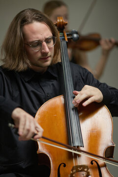 Vertical image of young adult Caucasian man playing cello during classical music orchestra rehearsal. Image suitable for arts education, concert advertising, symphonic performance promo