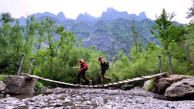 Two Hikers Crossing Wooden Suspension Bridge Over River in Mountain Forest