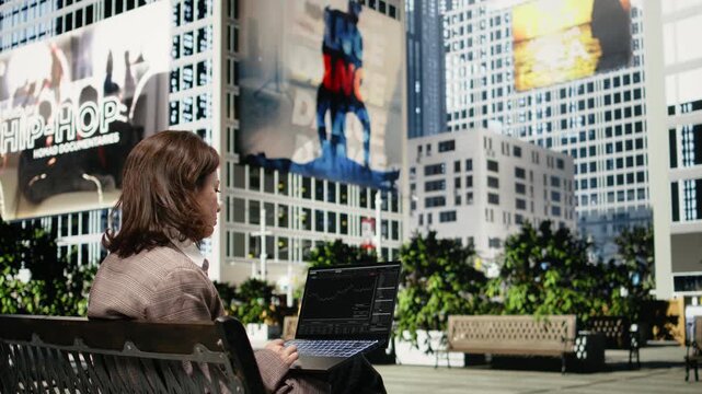 Trader reviews stock market charts on laptop seated on city park bench