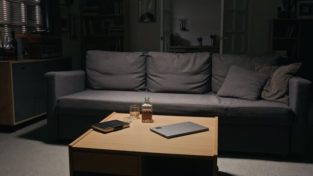 Alcohol bottle, drink glass, and laptop standing on table near empty couch in dim room. Scene suitable for alcohol abuse awareness, burnout, addiction, and mental wellbeing campaigns