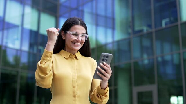 An excited businesswoman reading great news on a smartphone and celebrating success while standing near a modern glass office building. A happy female employee shocked by receiving a positive message.