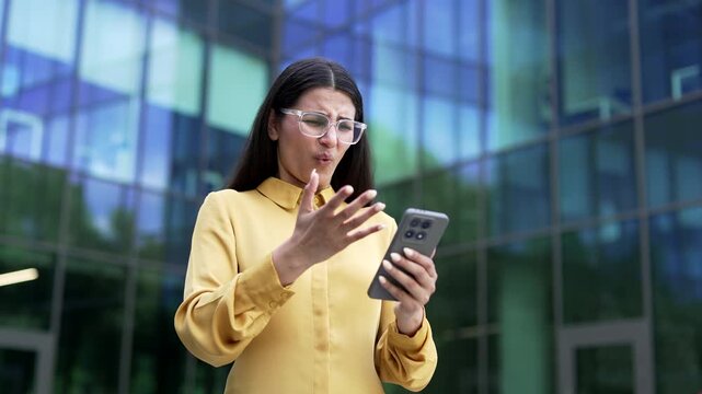 A confused businesswoman standing near a glass office building, struggling with an app error and declined mobile payment on a smartphone. A stressed female employee worried about an account access iss