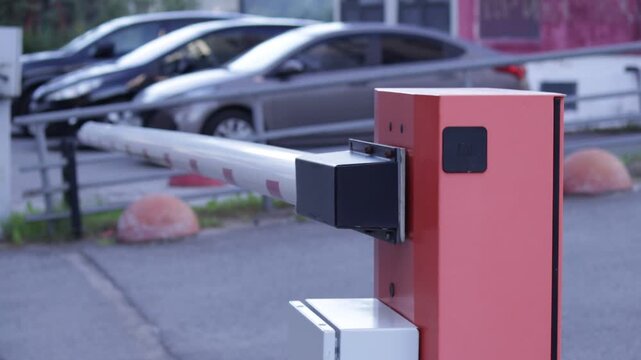 Close-up of a red automatic barrier post with white-and-red striped arm and black sensor module; blurred parked cars and building form background