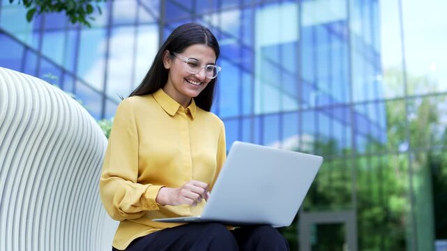 A smiling businesswoman reading good news on a laptop and feeling happy while sitting on a bench outdoors. A pleased female employee pleasantly surprised by a positive message near an office building.