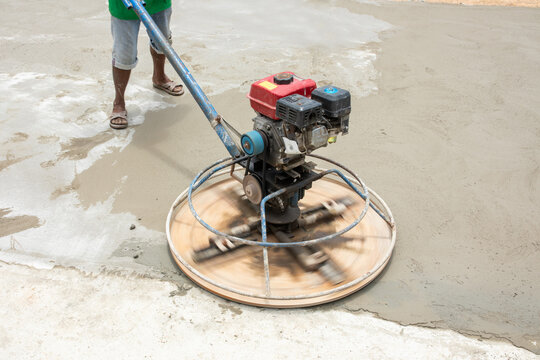 Power trowel disk spins in visible motion blur atop damp concrete floor, worker steps carefully behind machine, dynamic shot captures energy of rotation during finishing stage today