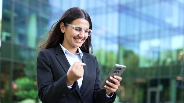 A close-up of an excited businesswoman in a formal suit, reading great news on a smartphone and celebrating success. A happy female employee shocked by a positive message near a modern office building