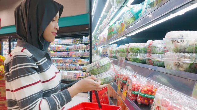 Asian Muslim woman selects fresh packaged produce while shopping in a brightly lit supermarket aisle, conveying healthy living, everyday errands, and mindful grocery shopping.