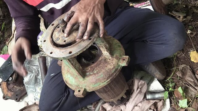 Greasy Hands striking a rusted metal circular flange coupling with a heavy hammer during an outdoor industrial water pump repair operation, framing concepts of rigorous manual labor and maintenance