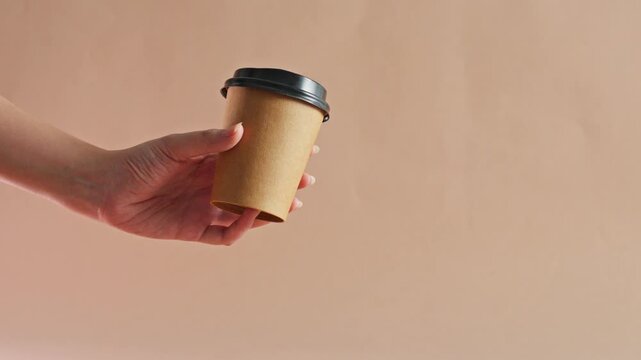 Hand holding eco-friendly brown paper coffee cup with lid against beige background