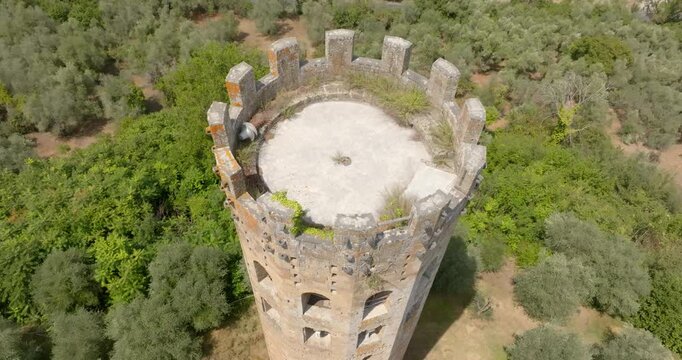 Aerial view of an ancient crenellated tower amid olive groves in the Italian countryside. The circular stone tower features arched window openings and a flat rooftop terrace.