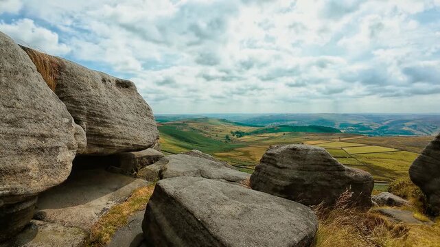 Granite rock formations with broad valley landscape under textured clouds slide shot. Patchwork fields between rolling hills and rugged stone outcrops. Rural countryside panorama with natural rock
