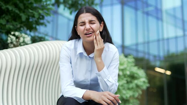 A businesswoman holding her cheek in pain while suffering from a severe toothache, sitting on a bench near an office building outdoors. A stressed female employee feeling tooth discomfort outside.