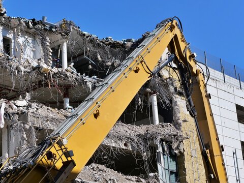 Yellow excavator long boom arm working at building demolition site, heavy machinery destroying concrete structure, urban redevelopment construction project