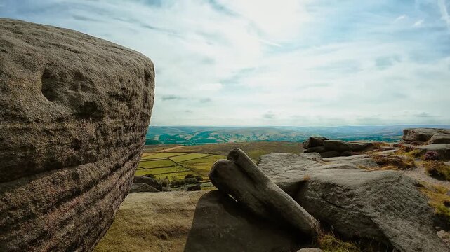 Weathered sandstone boulders on mountain ridge above green farmland and distant rolling hills. Rocky highland terrain beneath pale cloudy sky. Rural valley with fields and exposed stone