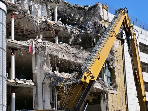 High reach yellow excavator arm dismantling damaged concrete building floors, urban redevelopment and heavy structural demolition site work
