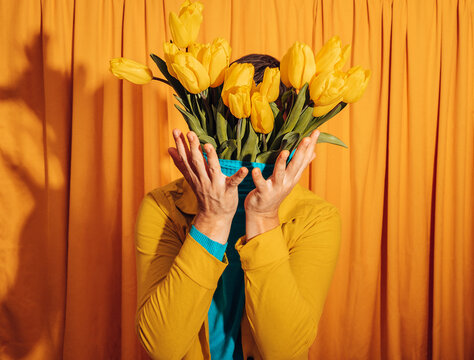 Man in yellow jacket hiding face with tulip vase in front of orange curtain