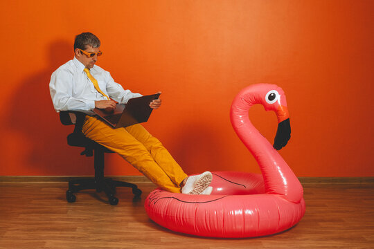 Tired man works on laptop with feet on inflatable flamingo indoors
