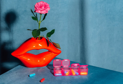 Red lip vase with a pink rose and a pillbox sit on a blue table