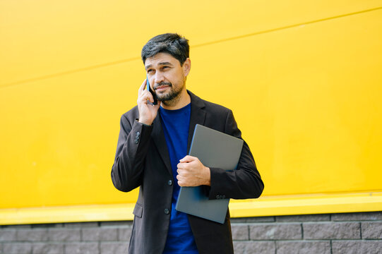 Businessman talks on phone holding laptop outdoors