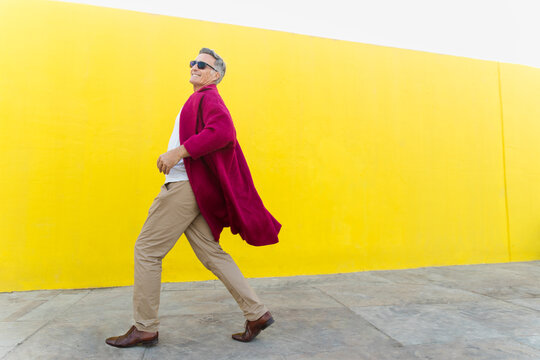 A man in a fuchsia coat walks happily past a yellow wall outdoors