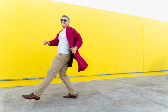 Happy man walking in fuchsia trench coat outdoors by yellow wall