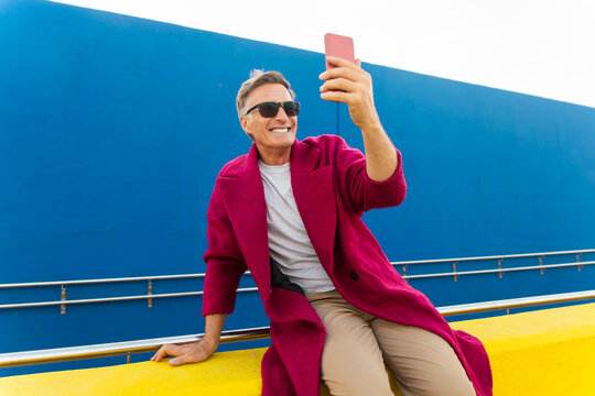 Man in fuchsia coat taking a selfie on a yellow wall