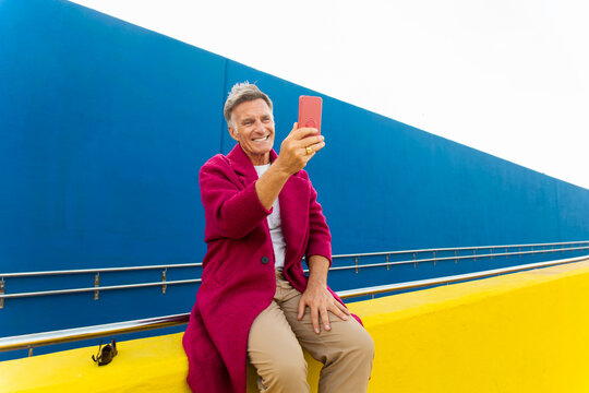 Smiling man in fuchsia coat takes selfie on yellow wall