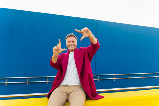 Smiling man in fuchsia coat makes a framing gesture outdoors