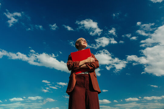 A smiling businesswoman holds a red folder against a blue sky