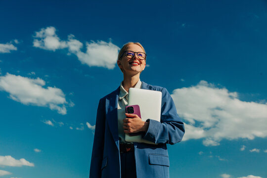 Smiling businesswoman holds laptop and phone outdoors under blue sky
