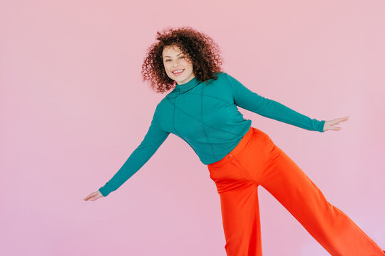 Smiling woman balancing playfully in colorful clothing on pink background