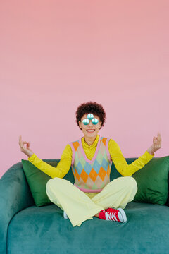 Smiling woman meditating on sofa with sunglasses and pink background