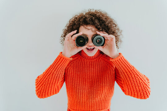 Curly haired woman in orange sweater smiling with binoculars indoors